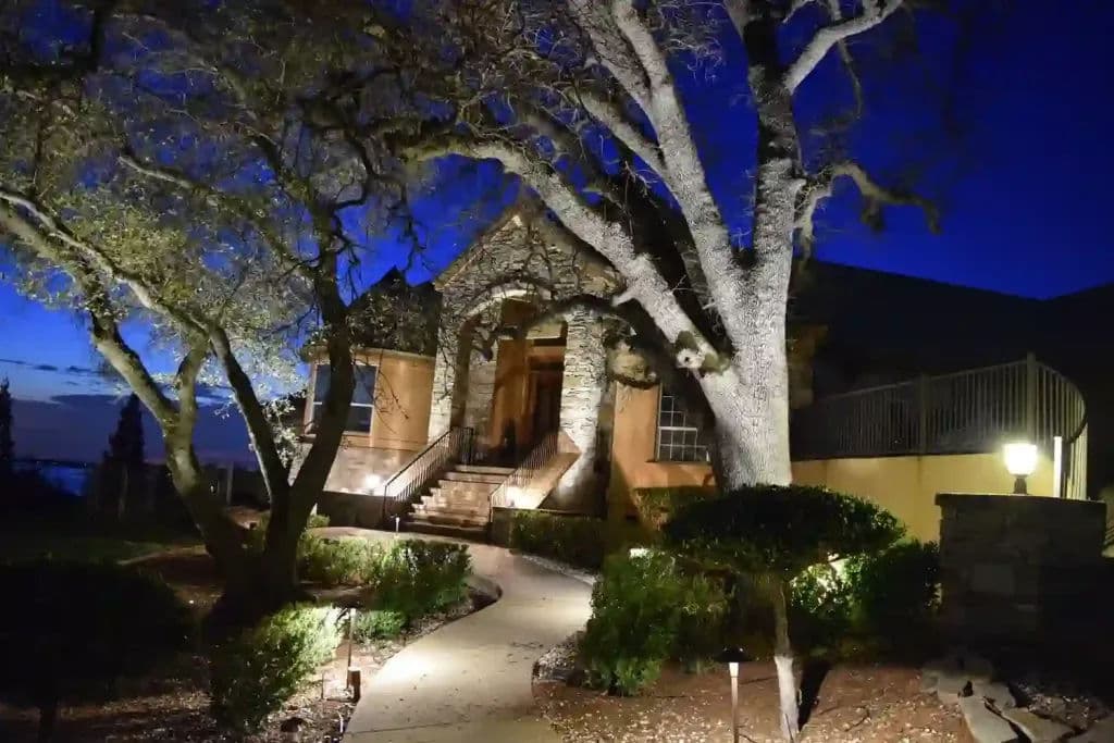 Elegant stone house at twilight, surrounded by trees and well-lit landscaping.