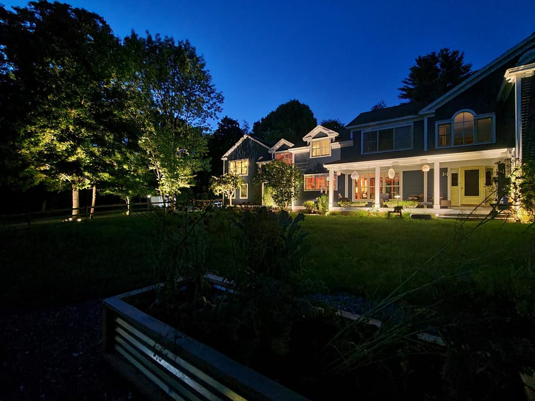 Twilight view of a beautifully lit house surrounded by trees and garden beds.