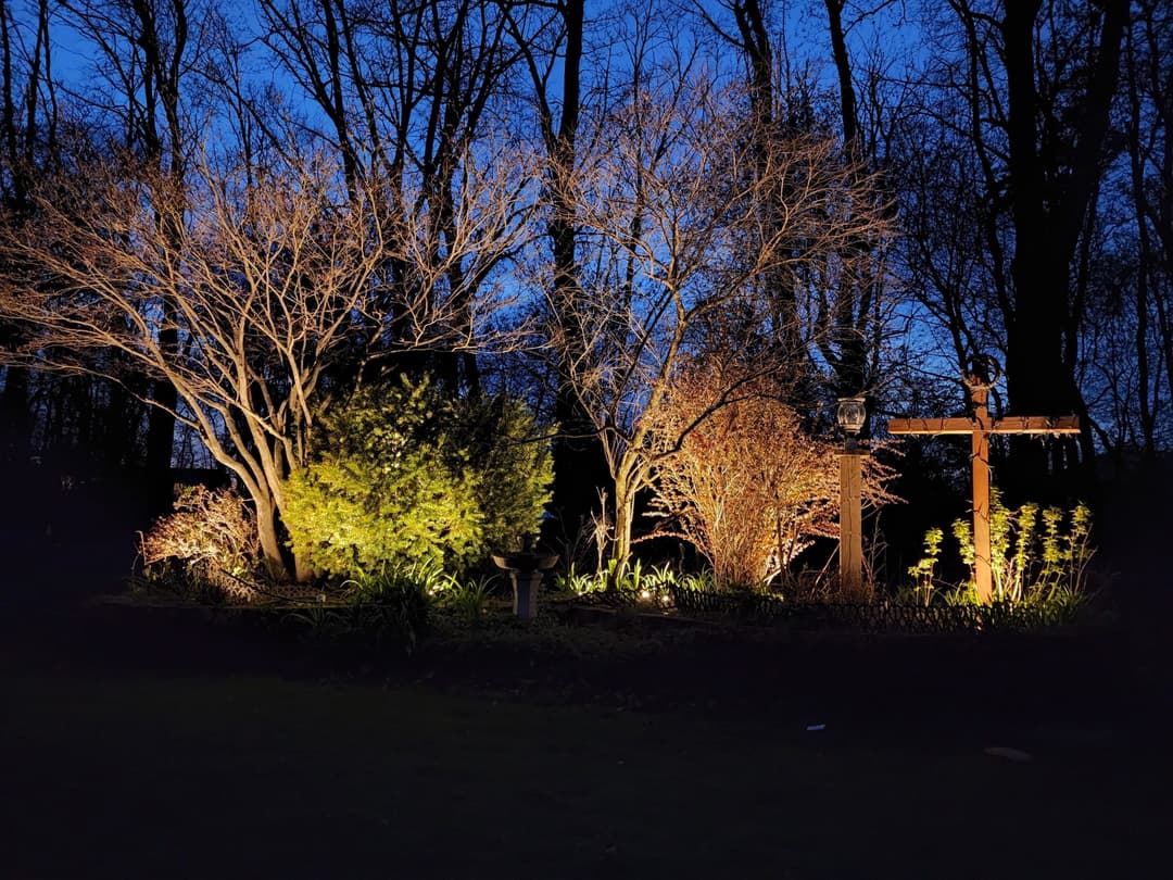 Illuminated garden landscape at dusk with trees and decorative cross among lush greenery.