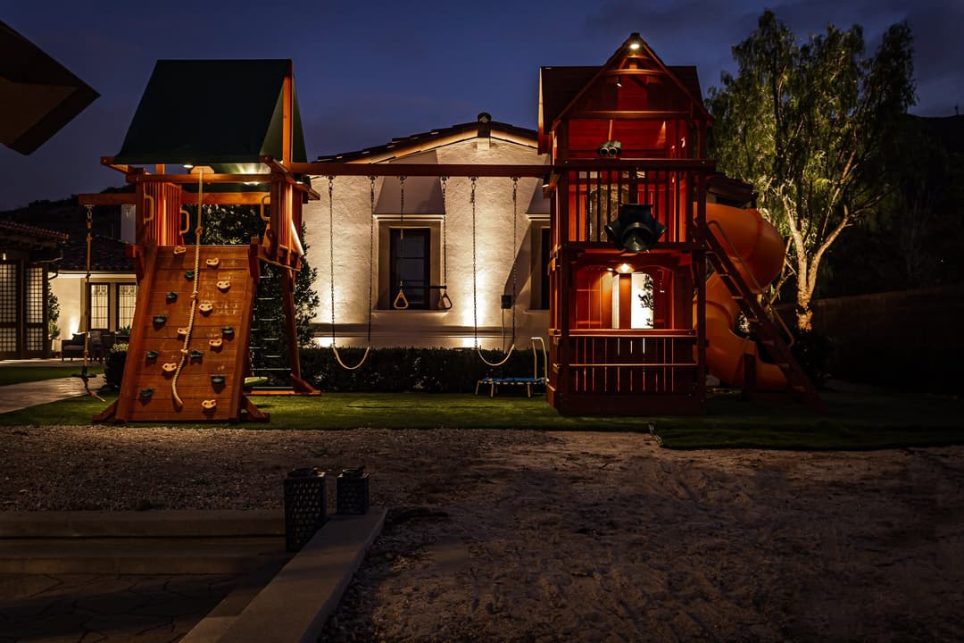 Playground at night with climbing structures and slides beside a house.