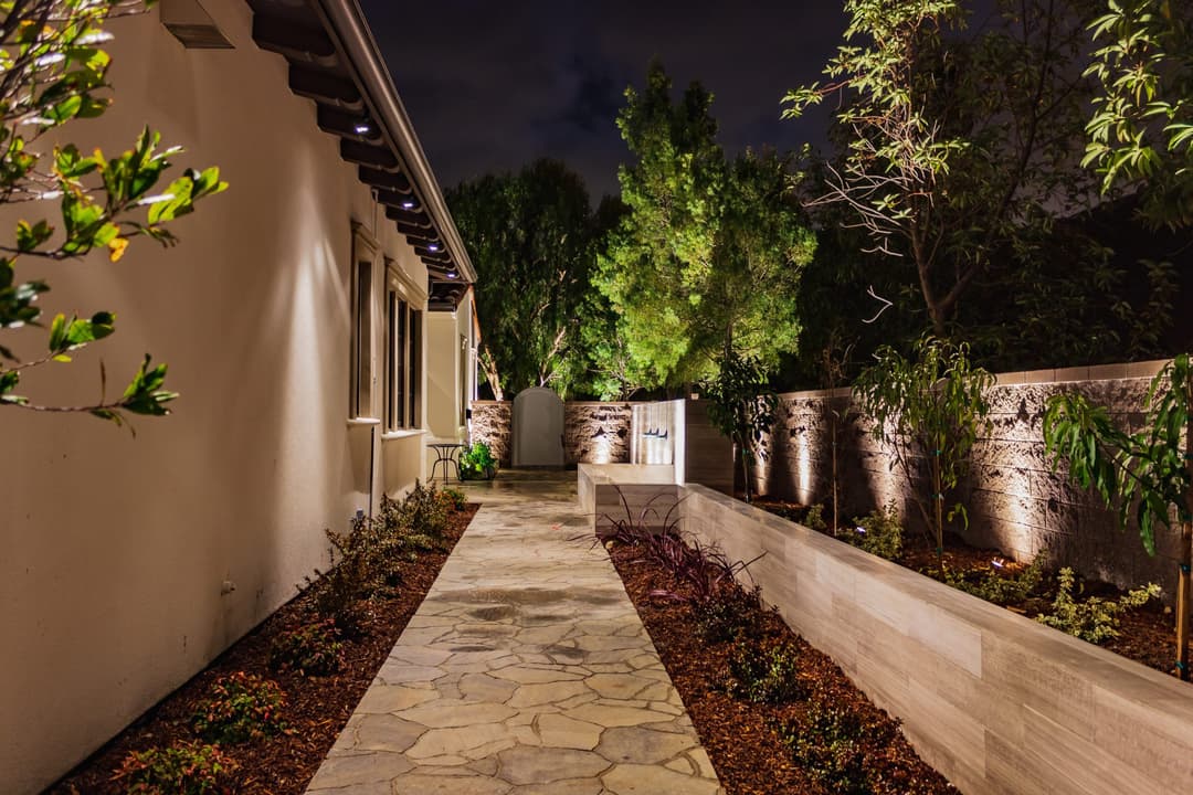 Well-lit garden pathway at night with stone walkway and lush greenery alongside a home.