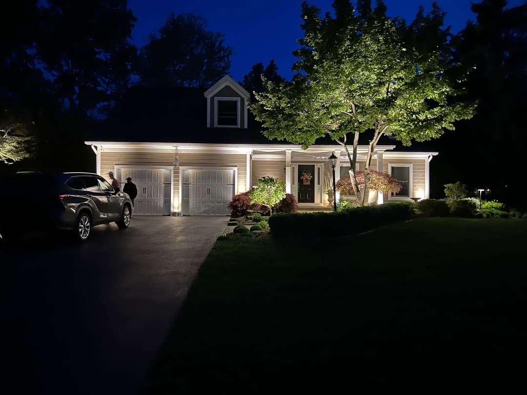 Illuminated suburban house at night with landscaped yard and parked car in driveway.