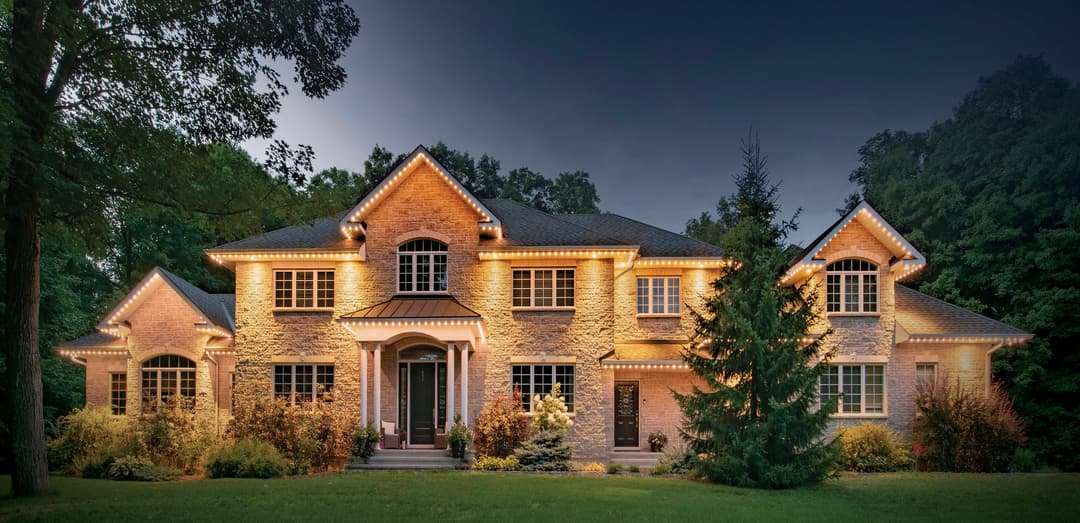 Elegant exterior of a two-story house illuminated with warm lights, surrounded by lush greenery.
