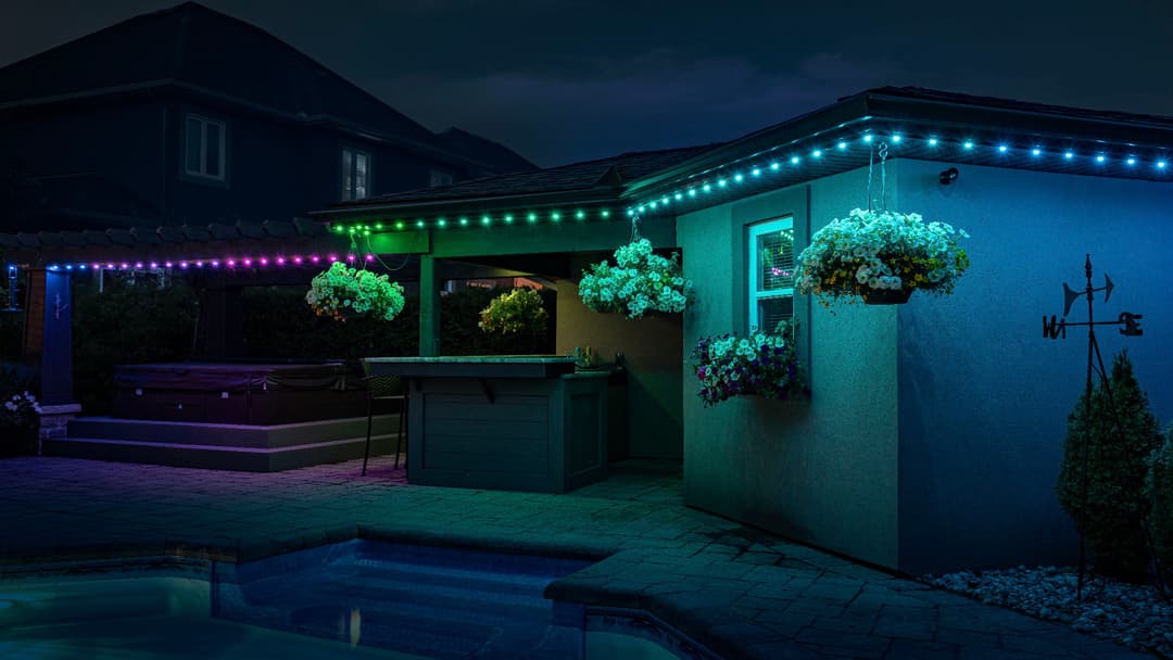 Colorful LED lights illuminate a patio with hanging flower baskets by a pool at night.
